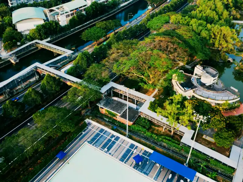 Vista aérea de uma área verde com estruturas urbanas, incluindo canais, passarelas e áreas de lazer, ao redor de árvores e vegetação.