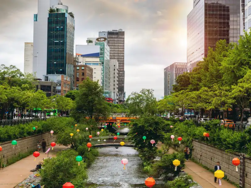 Vista da cidade com um rio decorado com lanternas coloridas, cercado por prédios altos e vegetação, criando um ambiente urbano encantador.